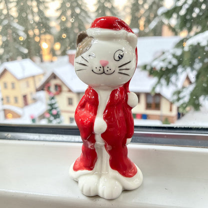 Keramikfigur einer Katze mit rotem Mantel und Weihnachtsmütze. Weiß-glänzende Oberfläche, stehende Position. Im Hintergrund ein Fenster mit winterlicher Szenerie und Häusern.