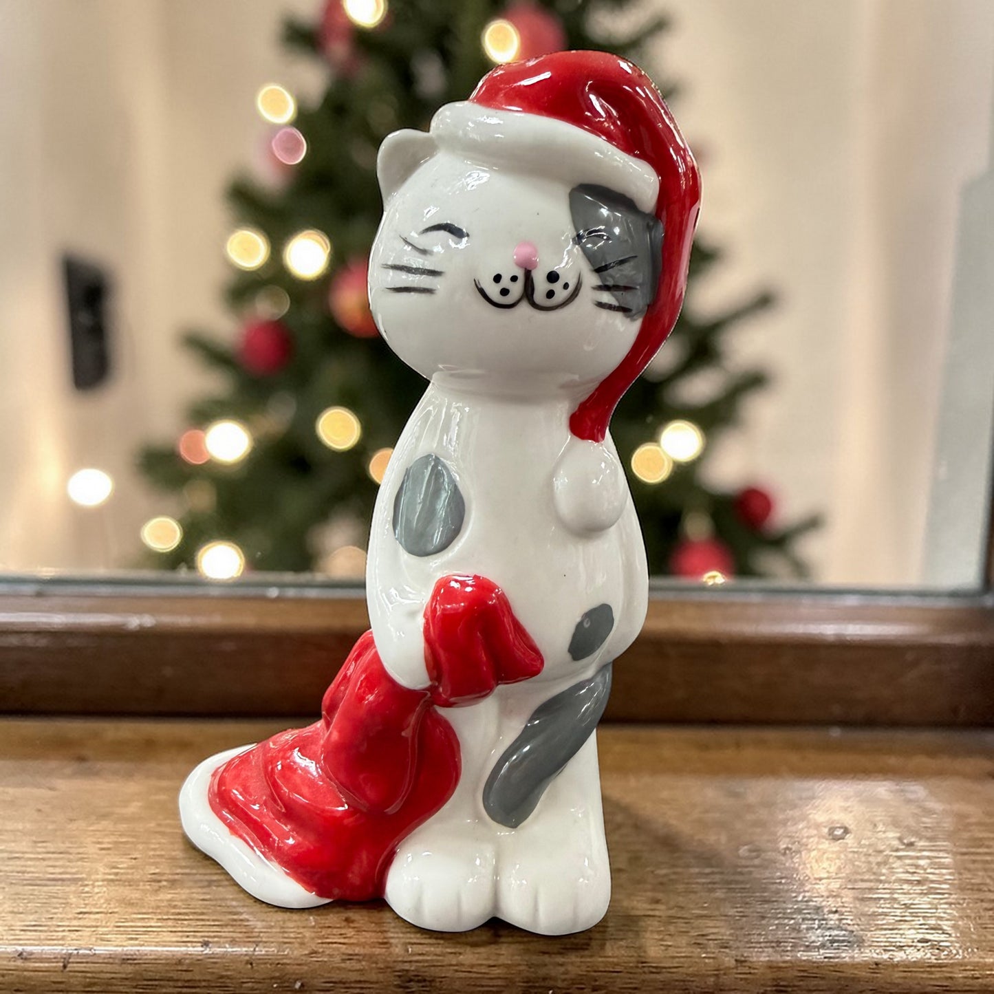 Keramikfigur einer Katze mit roter Weihnachtsmütze. Weiß mit grauen Flecken, stehende Haltung. Hintergrund mit unscharfem Weihnachtsbaum und Lichtpunkten.