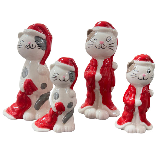 Vier Keramikfiguren in Form von Katzen in unterschiedlichen Größen. Weiß-glänzende Oberfläche mit grauen Details. Jede Figur trägt eine rote Weihnachtsmütze, zwei zusätzlich einen roten Mantel. Unterschiedliche Gesichtsausdrücke, teilweise mit zwinkerndem Auge. Freigestellt vor hellem Hintergrund.