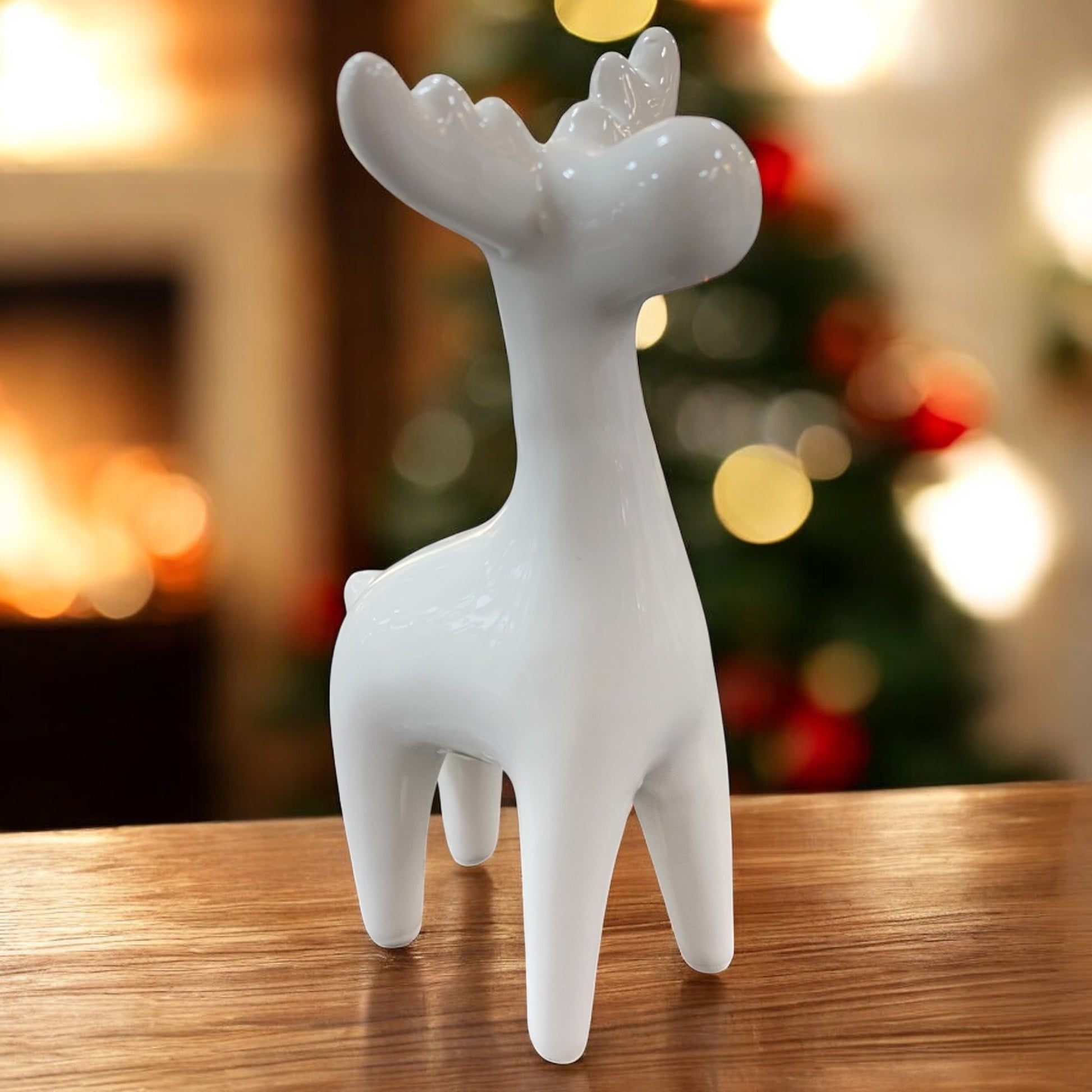Weiße Keramik-Elchfigur in modernem Stil auf einem Holztisch vor weihnachtlich beleuchtetem Hintergrund.