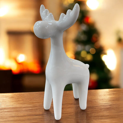Keramik-Elchfigur in Weiß auf einem Holztisch vor unscharfem Hintergrund mit Weihnachtsbaum und Kaminlicht.
