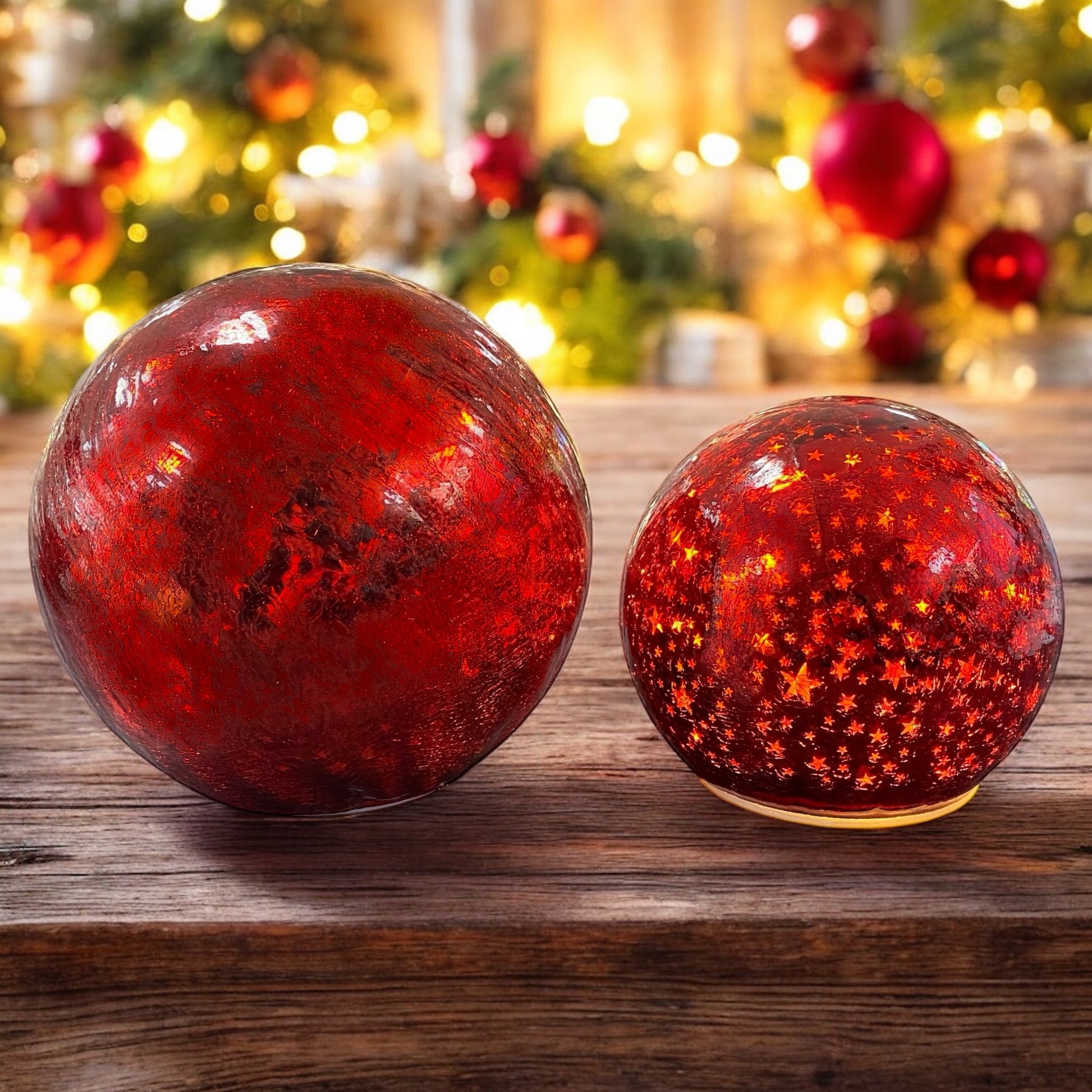 Zwei leuchtende rote Crackle-Glas-Kugeln mit goldenen Sternen auf einem Holztisch vor warmem Lichterhintergrund.