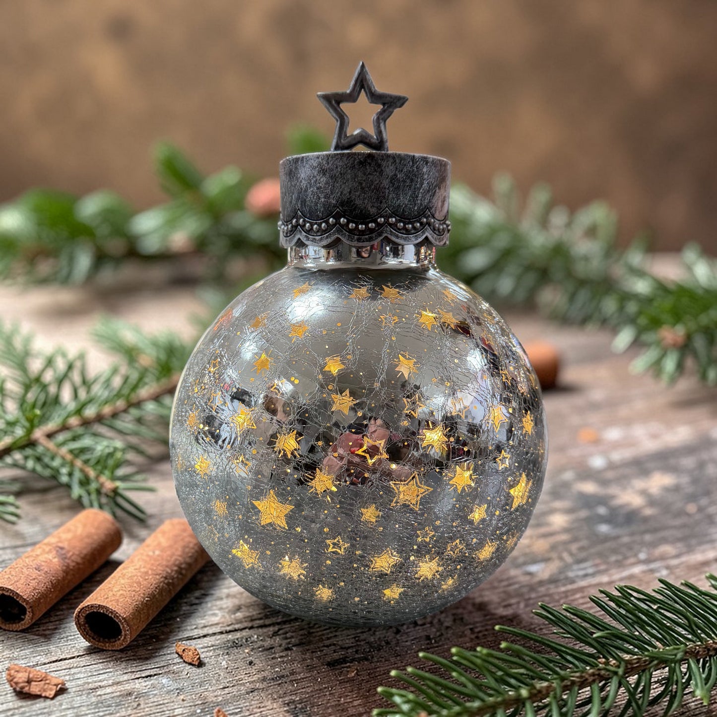 Stimmungsvolle Weihnachtsdekoration mit silberner Glaskugel und goldenen Sternen, arrangiert auf einer rustikalen Holzfläche mit Tannenzweigen und Zimtstangen. Die Kombination aus natürlichen Elementen und glänzendem Glas schafft ein warmes, festliches Ambiente.