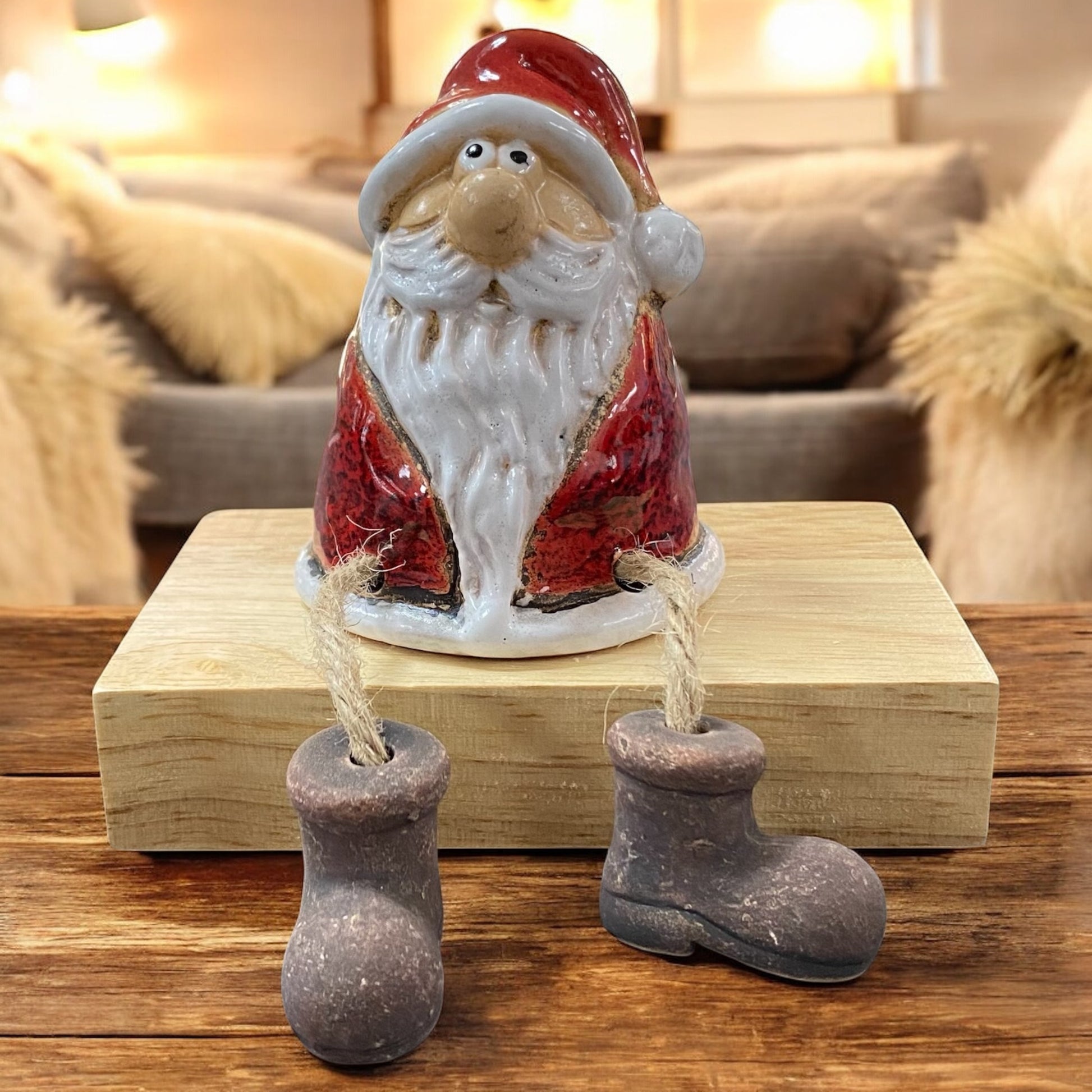Keramik-Weihnachtsmann mit rotem Mantel und langen Beinen als Kantenhocker auf einem Holzblock vor gemütlichem Wohnzimmerhintergrund.

