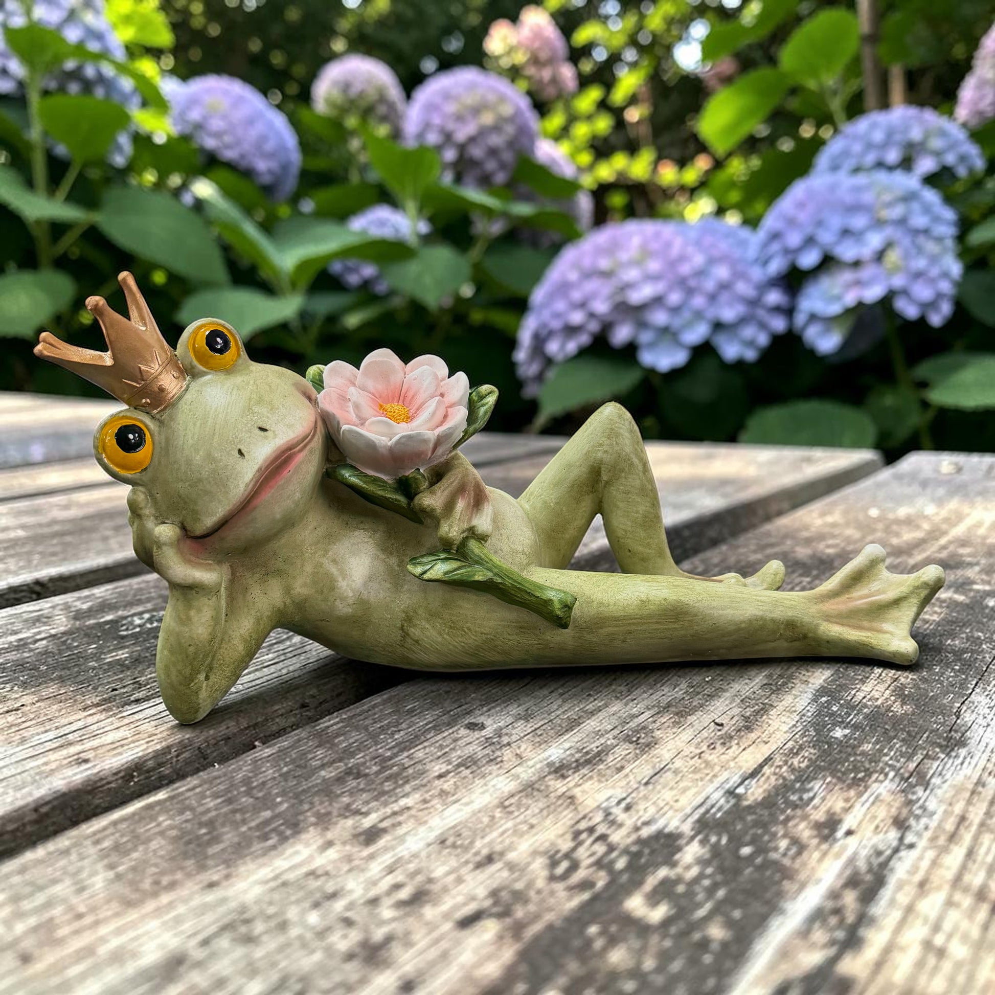 Grüne Froschkönig-Dekofigur mit goldener Krone und rosa Blüte auf einem Holztisch vor blühenden Hortensien.