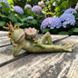 Grüne Froschkönig-Dekofigur mit goldener Krone und rosa Blüte auf einem Holztisch vor blühenden Hortensien.
