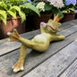 Grüne Froschkönig-Dekofigur mit goldener Krone und rosa Tulpe auf einem Holztisch vor grünen Pflanzen.