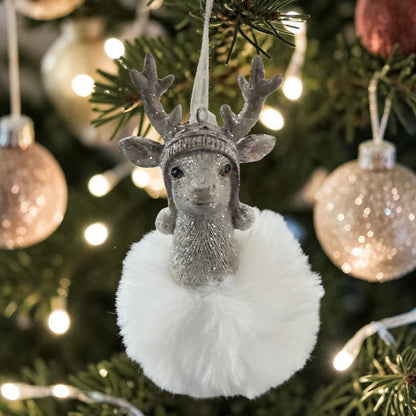 Hängedekoration mit Rentierfigur in silberfarbener Oberfläche und weißer Plüschkugel, aufgehängt an einem grünen Weihnachtsbaum mit warmen Lichtern und goldfarbenen Kugeln im Hintergrund.