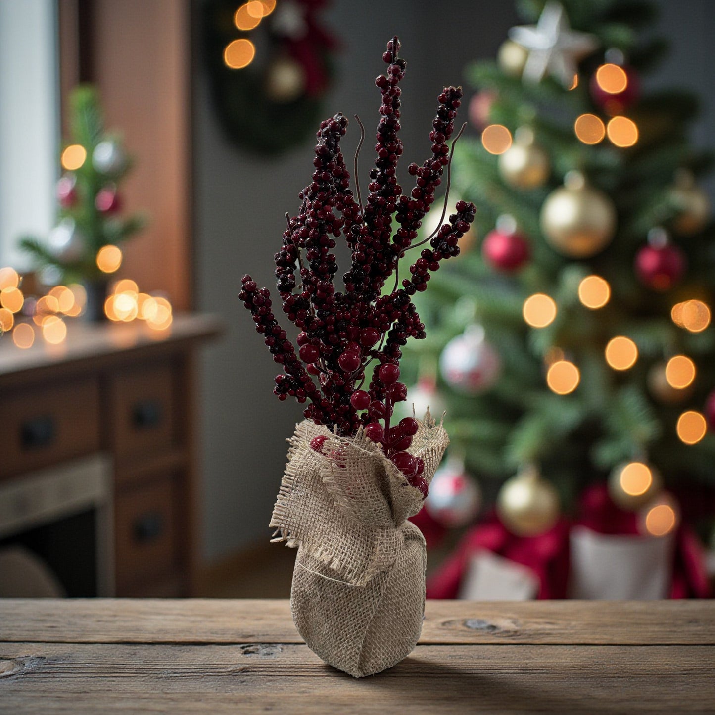 Ein Arrangement aus roten Beerenzweigen in einem Jutesack steht auf einer Holzfläche vor einem festlich dekorierten Weihnachtsbaum mit warmem Licht.