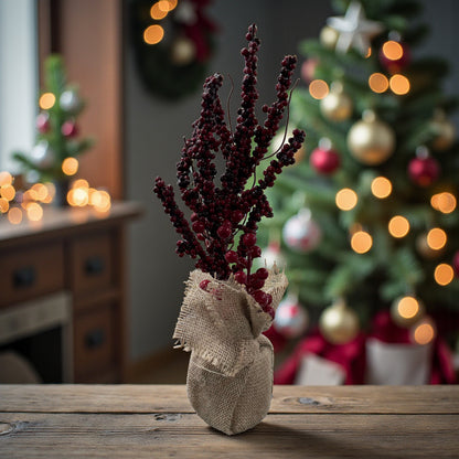 Ein Arrangement aus roten Beerenzweigen in einem Jutesack steht auf einer Holzfläche vor einem festlich dekorierten Weihnachtsbaum mit warmem Licht.
