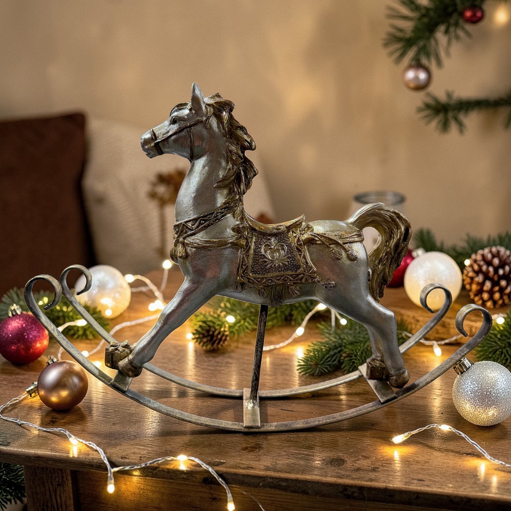 Ein einzelnes Schaukelpferd in gold-champagnerfarbenem Vintage-Design steht dekorativ auf einem Tisch mit Weihnachtskugeln, Tannenzweigen und Lichterkette.