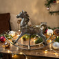 Ein einzelnes Schaukelpferd in gold-champagnerfarbenem Vintage-Design steht dekorativ auf einem Tisch mit Weihnachtskugeln, Tannenzweigen und Lichterkette.