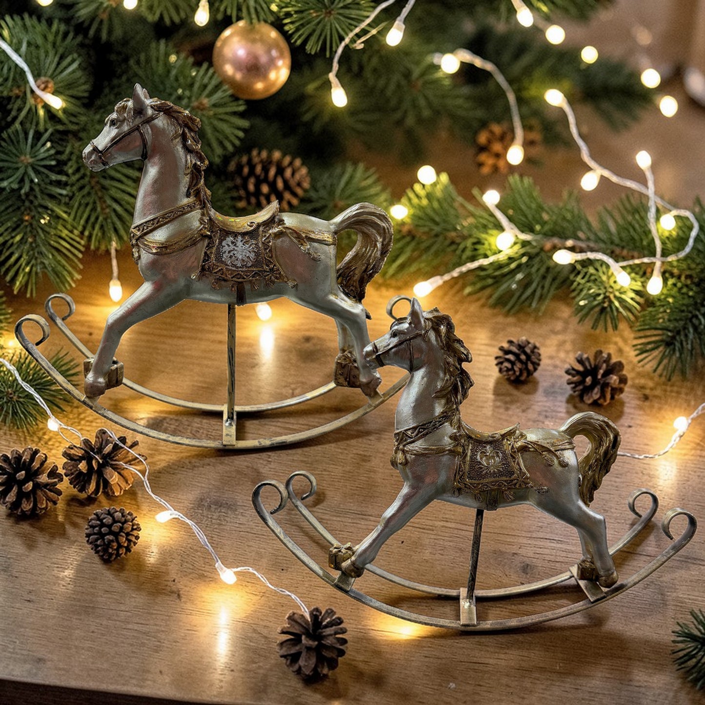 Zwei unterschiedlich große Schaukelpferde in gold-champagnerfarbenem Look stehen auf einem Holztisch, umgeben von Tannenzweigen, Zapfen und einer warm leuchtenden Lichterkette.