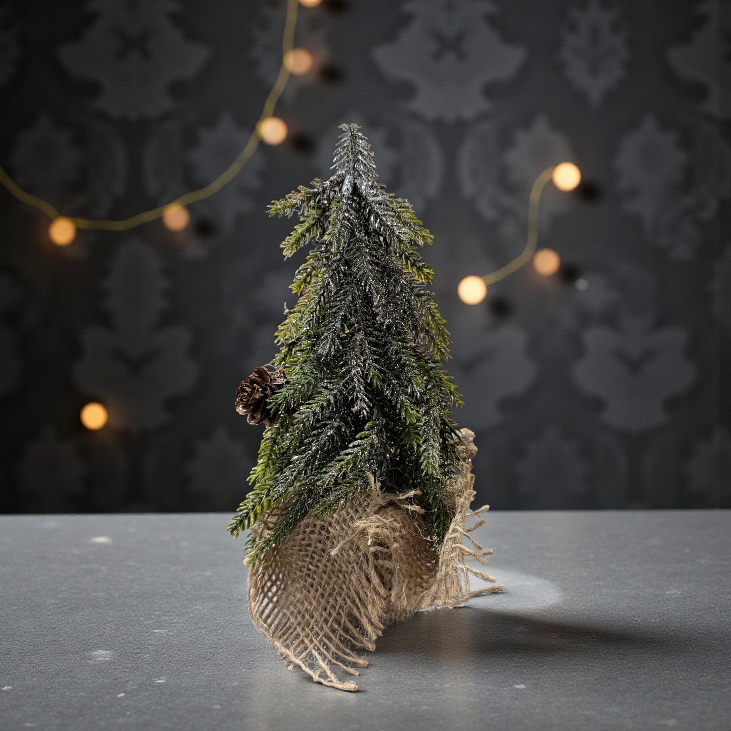 Ein einzelner grüner Dekotannenbaum mit Zapfen in einem Jutebeutel steht auf einer dunklen Oberfläche vor einem dekorativen Hintergrund mit Lichterkette.