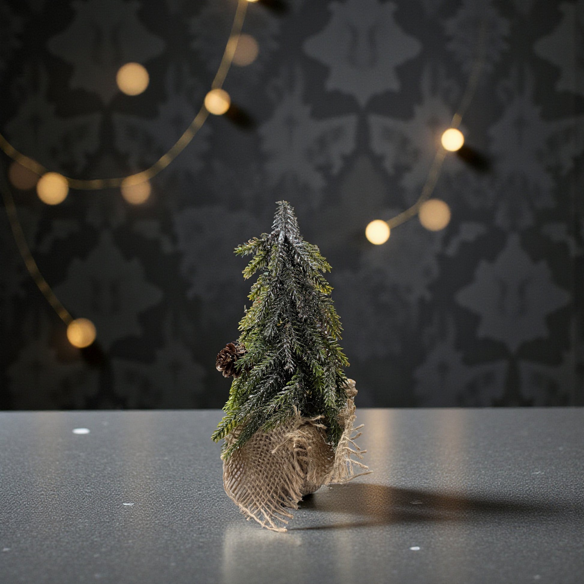 Ein einzelner kleiner grüner Dekotannenbaum mit Zapfen in einem Jutebeutel steht auf einer dunklen Fläche vor einer dezenten Lichterkette.