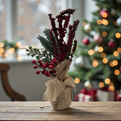 Ein Arrangement aus roten Beerenzweigen und grünen Tannenzweigen in einem Jutesack steht auf einer Holzoberfläche vor einem Fenster und einem geschmückten Weihnachtsbaum.