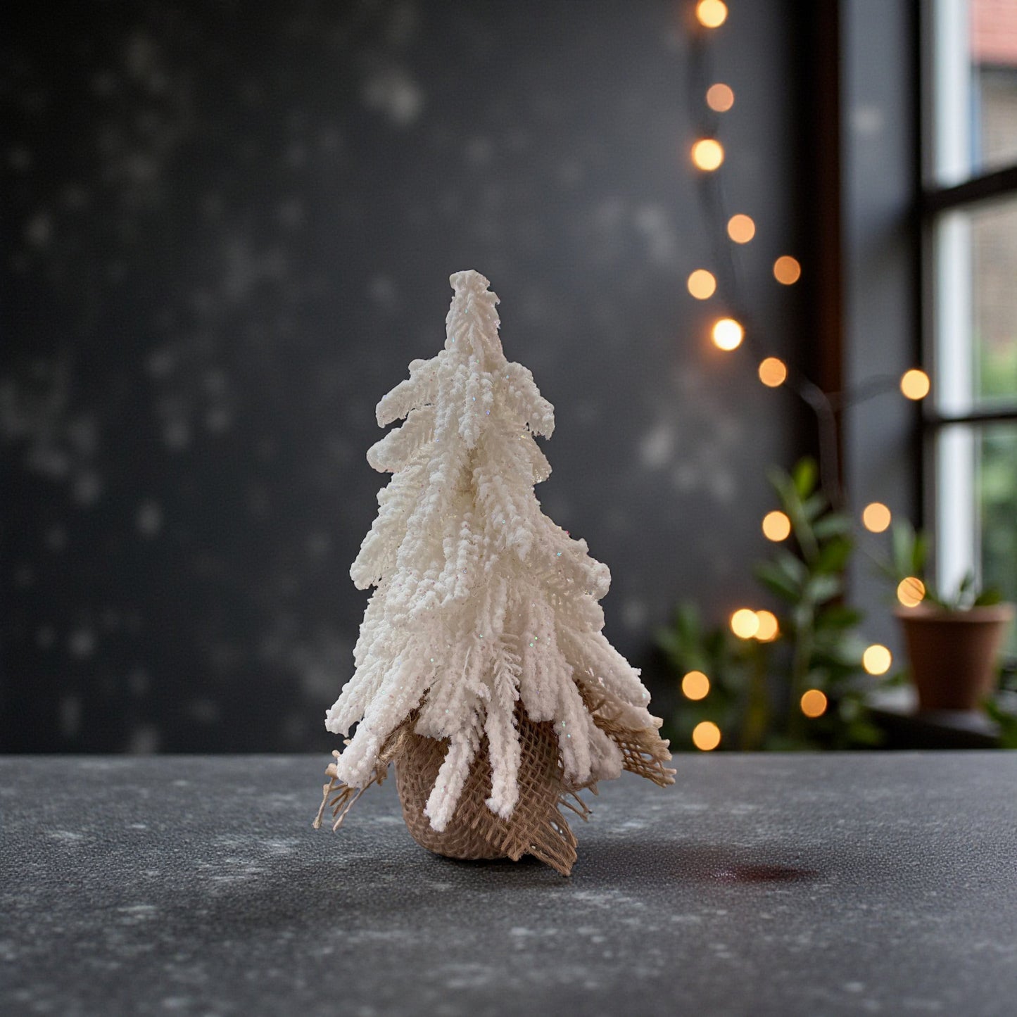 Ein einzelner kleiner, weißer Dekotannenbaum in einem Jutebeutel steht auf einer dunklen Fläche vor einem Fenster und einer dezent beleuchteten Lichterkette.