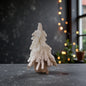 Ein einzelner kleiner, weißer Dekotannenbaum in einem Jutebeutel steht auf einer dunklen Fläche vor einem Fenster und einer dezent beleuchteten Lichterkette.