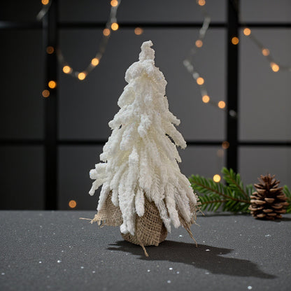 Ein einzelner weißer Dekotannenbaum in einem Jutebeutel steht auf einer dunklen Oberfläche, daneben liegen ein Tannenzweig und ein Tannenzapfen vor einem Hintergrund mit Lichterkette.