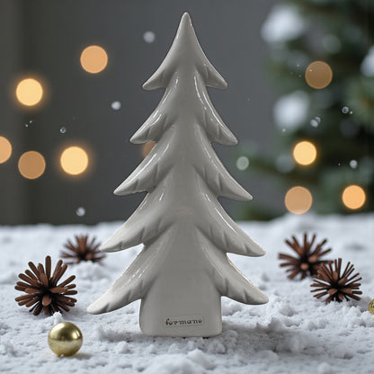 Ein cremeweißer Keramik-Tannenbaum steht auf einer schneebedeckten Oberfläche, umgeben von Zapfen und goldenen Kugeln vor einem weich beleuchteten Hintergrund.