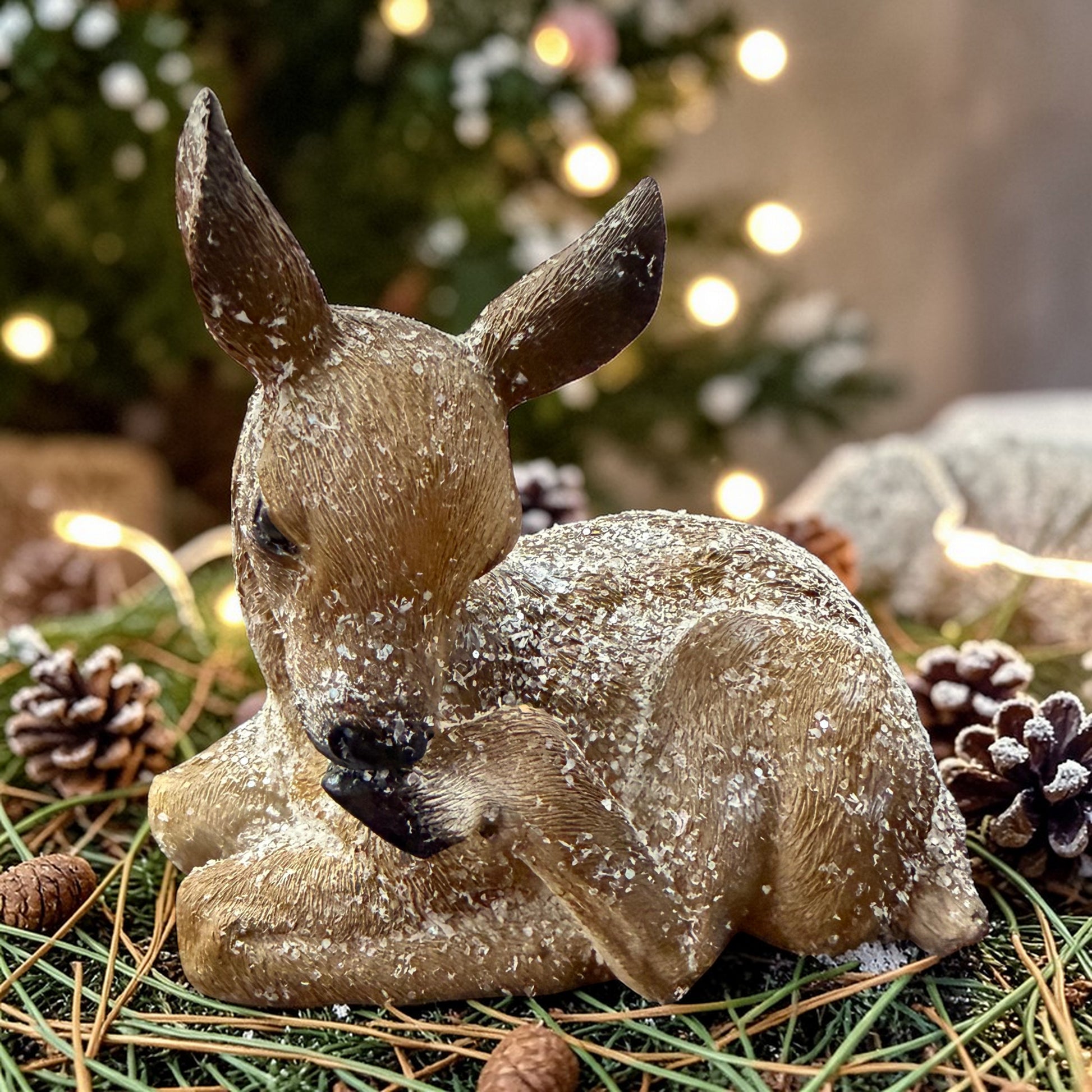 Ein liegendes Reh mit realistischer Fellstruktur und Kunstschnee liegt dekorativ auf Tannenzweigen mit Zapfen vor warmen Lichtern im Hintergrund.