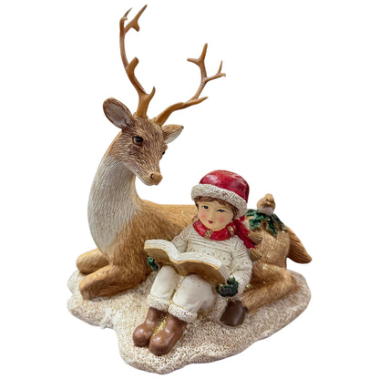 Eine dekorative Figur zeigt ein sitzendes Kind mit roter Mütze, das ein Buch liest, neben einem Hirsch und einem Reh auf einer winterlichen Basis.