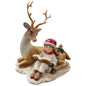 Eine dekorative Figur zeigt ein sitzendes Kind mit roter Mütze, das ein Buch liest, neben einem Hirsch und einem Reh auf einer winterlichen Basis.