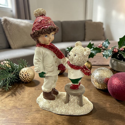 Eine dekorative Figur zeigt ein Kind, das einem Teddybären auf einem Hocker gegenübersteht.