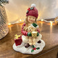 Eine sitzende Mädchenfigur mit roter Mütze hält einen Teddy und eine Tasse in den Händen und sitzt auf einer winterlich gestalteten Basis.