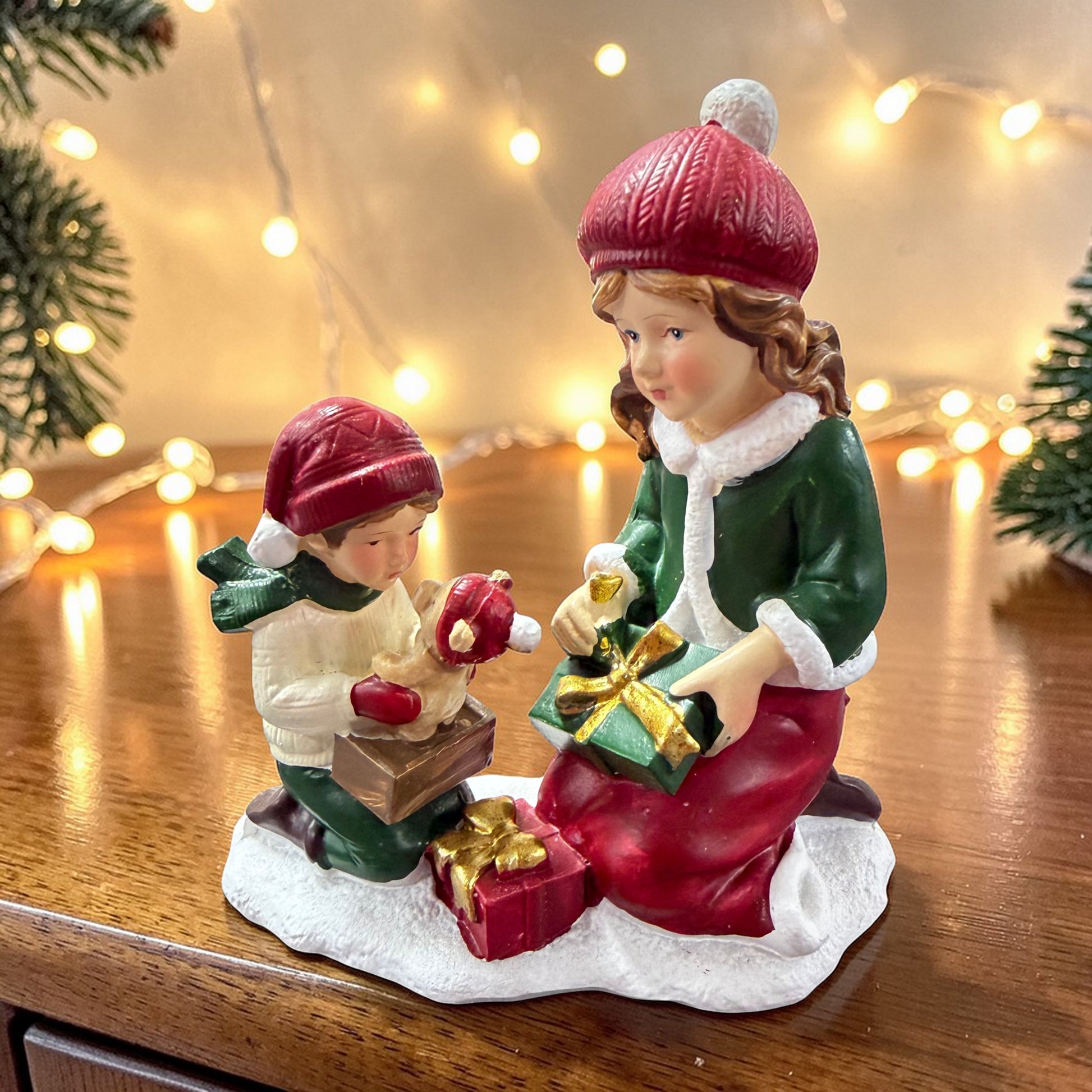 Zwei sitzende Kinderfiguren mit roten Mützen sind beim Auspacken von Geschenken dargestellt und befinden sich auf einer winterlichen Standfläche.