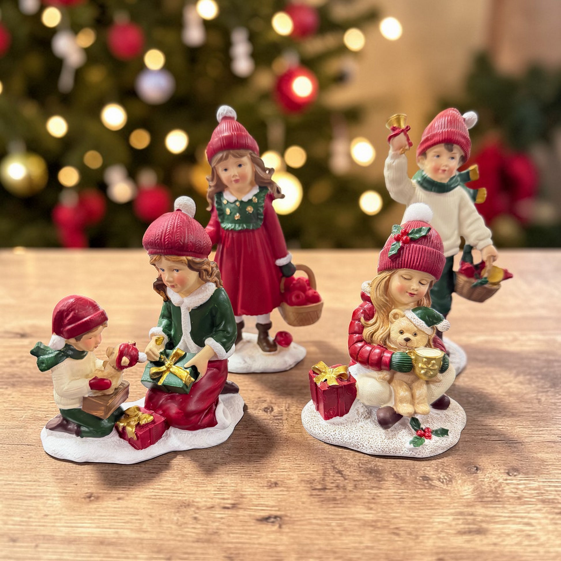 Mehrere dekorative Kinderfiguren mit roten Mützen stehen auf einer Holzoberfläche vor einem unscharfen Weihnachtsbaum mit Lichterdekoration.