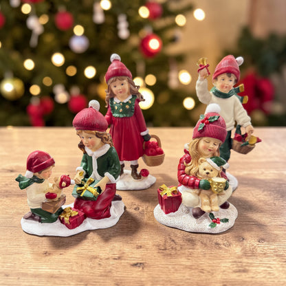 Mehrere dekorative Kinderfiguren mit roten Mützen stehen auf einer Holzoberfläche vor einem unscharfen Weihnachtsbaum mit Lichterdekoration.