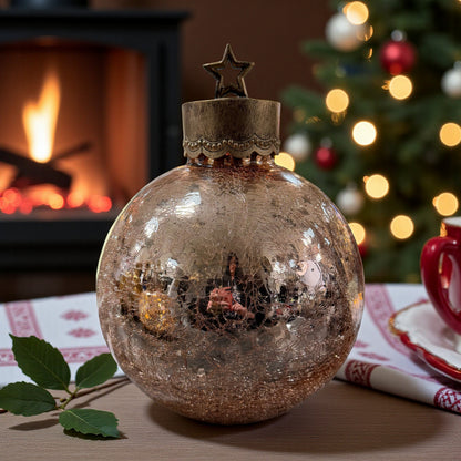 Eine kugelförmige Dekoleuchte aus crackle Glas mit Sternmuster und Metallkappe mit Stern steht auf einem Tisch vor einem Kamin und einem geschmückten Weihnachtsbaum.