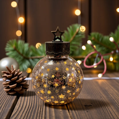 Eine kleine kugelförmige Dekoleuchte aus crackle Glas mit Sternmuster und Metallkappe mit Stern steht auf einem Holztisch vor Tannenzweigen und einer Lichterkette.