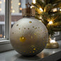 Eine taupefarbene Dekokugel mit Stern- und Sternschnuppenmuster steht auf einer Fensterbank neben einem geschmückten Weihnachtsbaum.