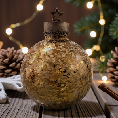Eine kugelförmige Dekoleuchte aus Glas mit eingraviertem Tannenmotiv und Metallkappe mit Stern steht auf einem Holztisch neben Zapfen und Lichterkette.