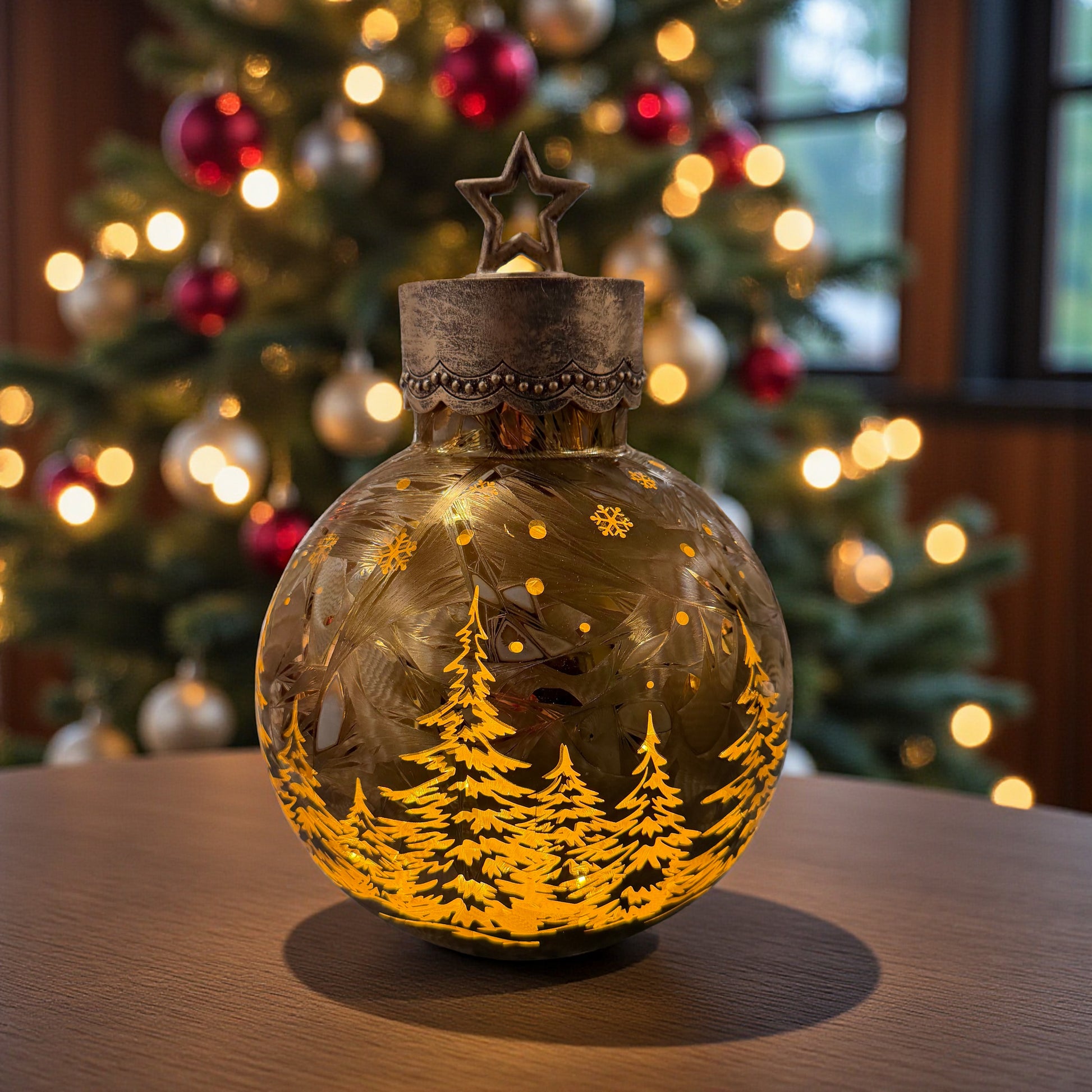 Eine kugelförmige Dekoleuchte aus Glas mit eingraviertem Tannenmotiv und Metallkappe mit Stern steht auf einem Tisch vor einem geschmückten Weihnachtsbaum.