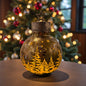 Eine kugelförmige Dekoleuchte aus Glas mit eingraviertem Tannenmotiv und Metallkappe mit Stern steht auf einem Tisch vor einem geschmückten Weihnachtsbaum.