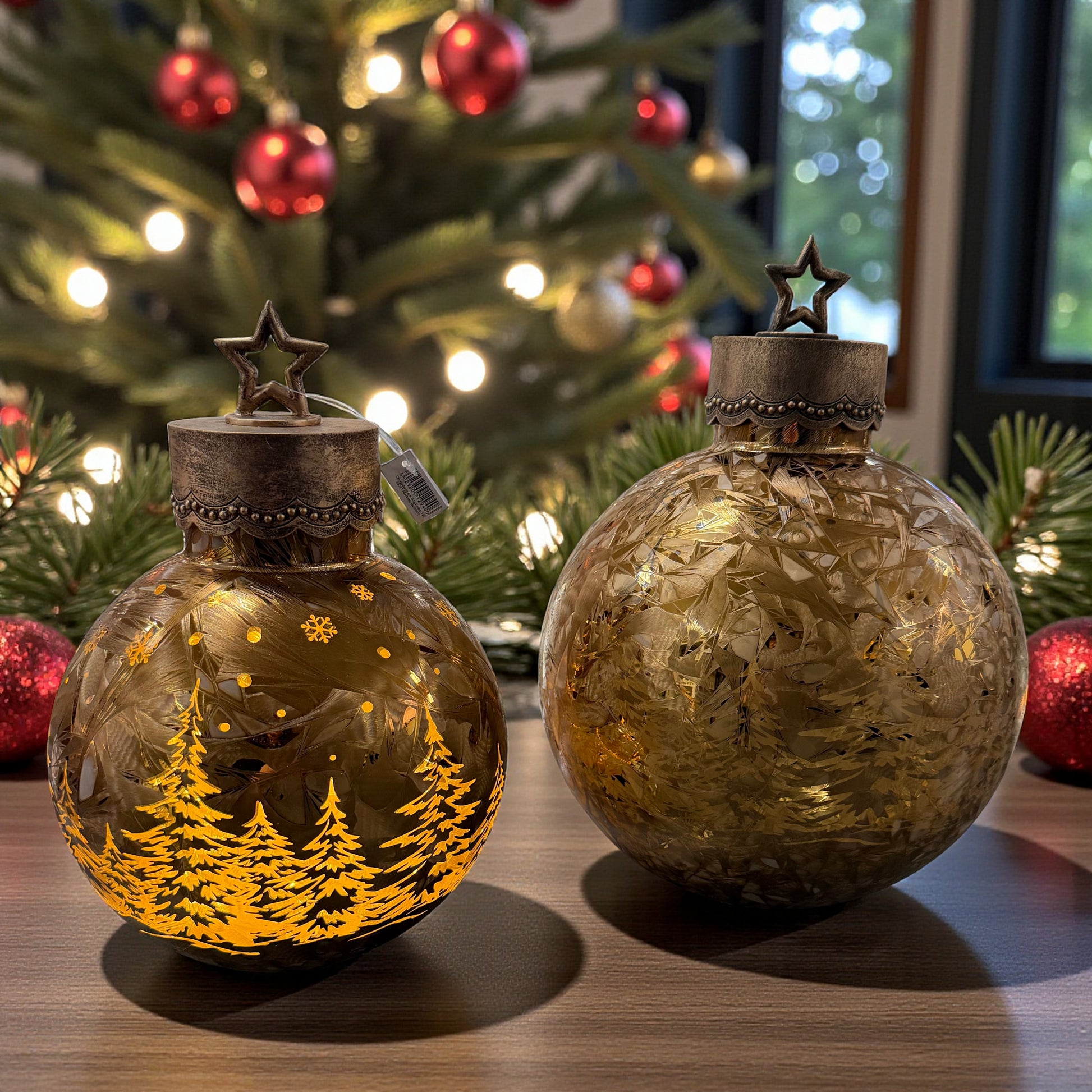 Zwei kugelförmige Dekoleuchten aus Glas mit eingraviertem Tannenmotiv und Metallkappen mit Stern stehen auf einem Tisch vor einem geschmückten Weihnachtsbaum.