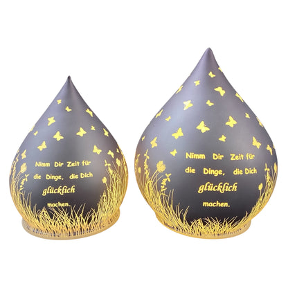 Zwei lila-farbene Dekolicht Tropfen aus Glas mit Spruch "Nimm dir Zeit für die Dinge, die dich glücklich machen" vor weißem Hintergrund