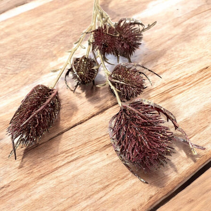 Künstlicher Distelzweig in Bordeaux und Gold mit mehreren glitzernden Blütenköpfen und feinen, verzweigten Stielen auf hölzernem Hintergrund