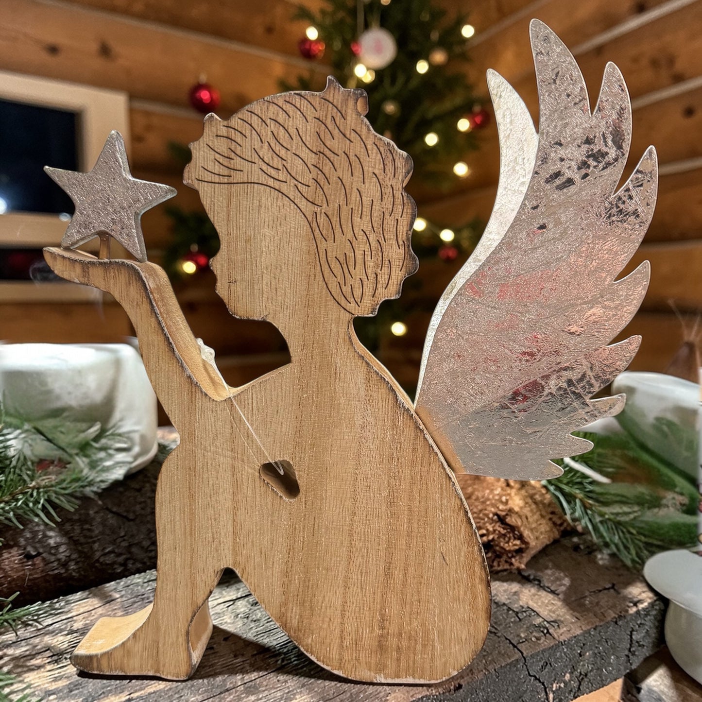 Holzfigur in Form eines Engels mit silberfarbenen Flügeln und einem Stern in der Hand auf einer dekorierten Oberfläche vor weihnachtlichem Hintergrund.