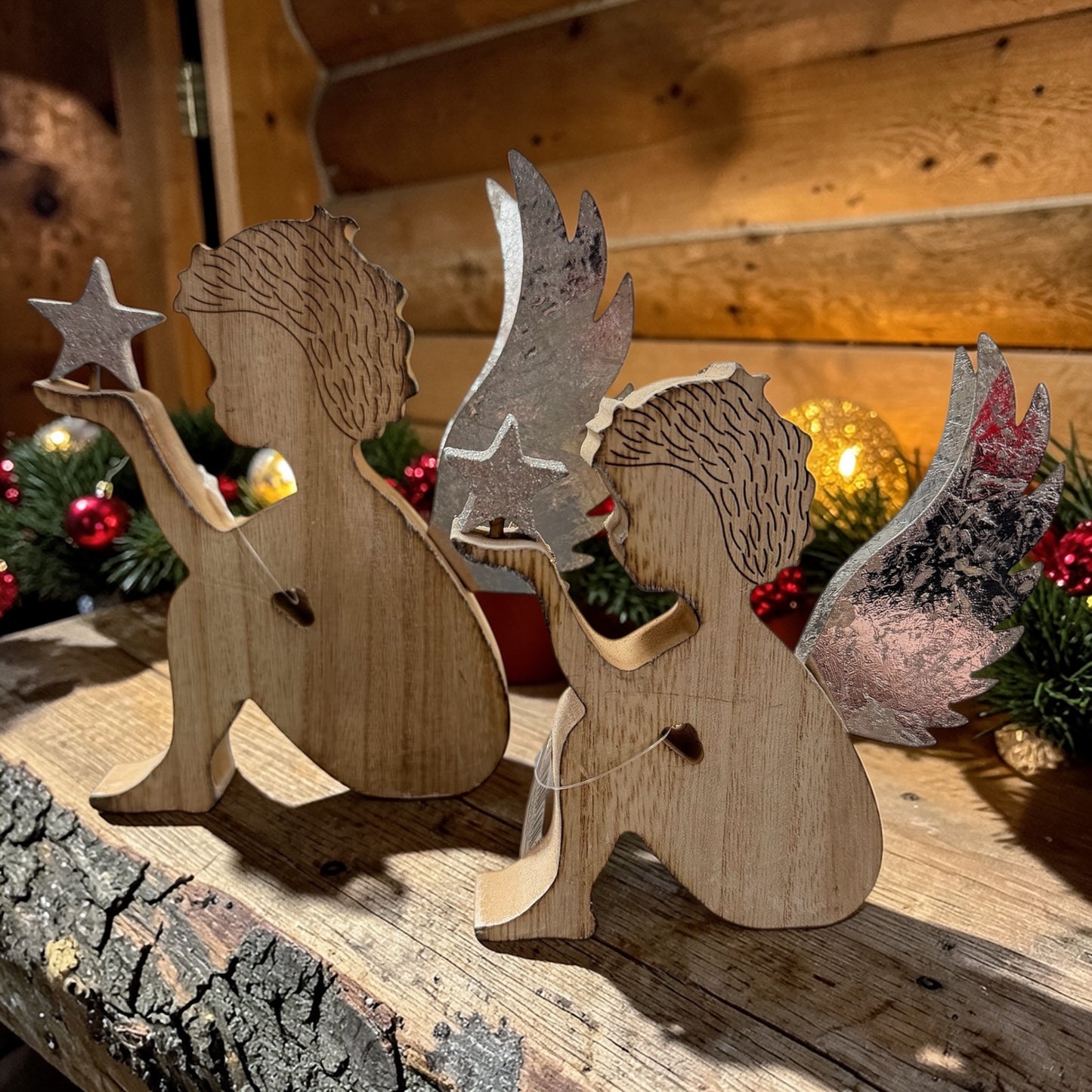 Zwei Holzfiguren in Form von Engeln mit silberfarbenen Flügeln auf einer Holzfläche mit weihnachtlicher Dekoration im Hintergrund.