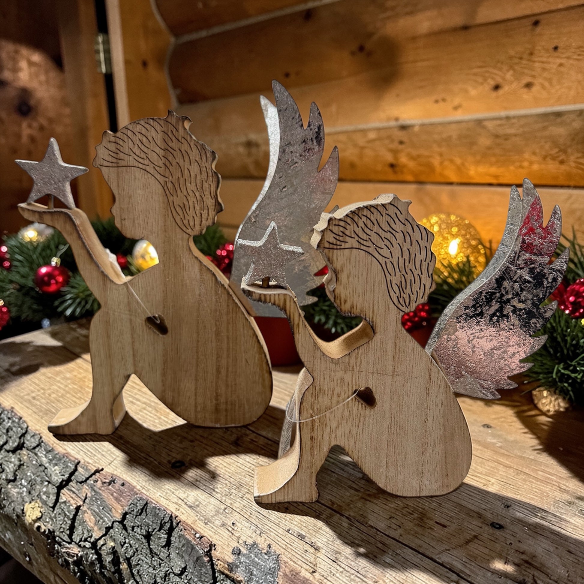 Zwei Holzfiguren in Form von Engeln mit silberfarbenen Flügeln auf einer Holzfläche mit weihnachtlicher Dekoration im Hintergrund.