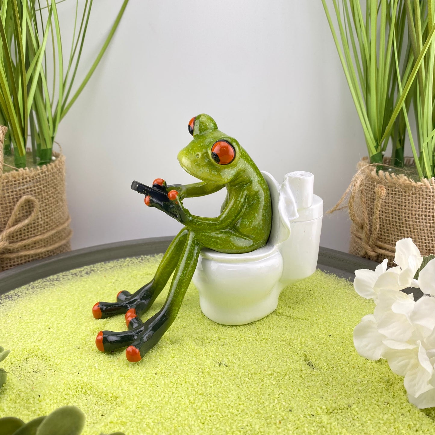 Eine Dekofigur im Ambiente, die einen Frosch auf Toilette abbildet. Der Deko Frosch ist einer ganz alltäglichen Situation mit seinem Smartphone in der Hand dargestellt.