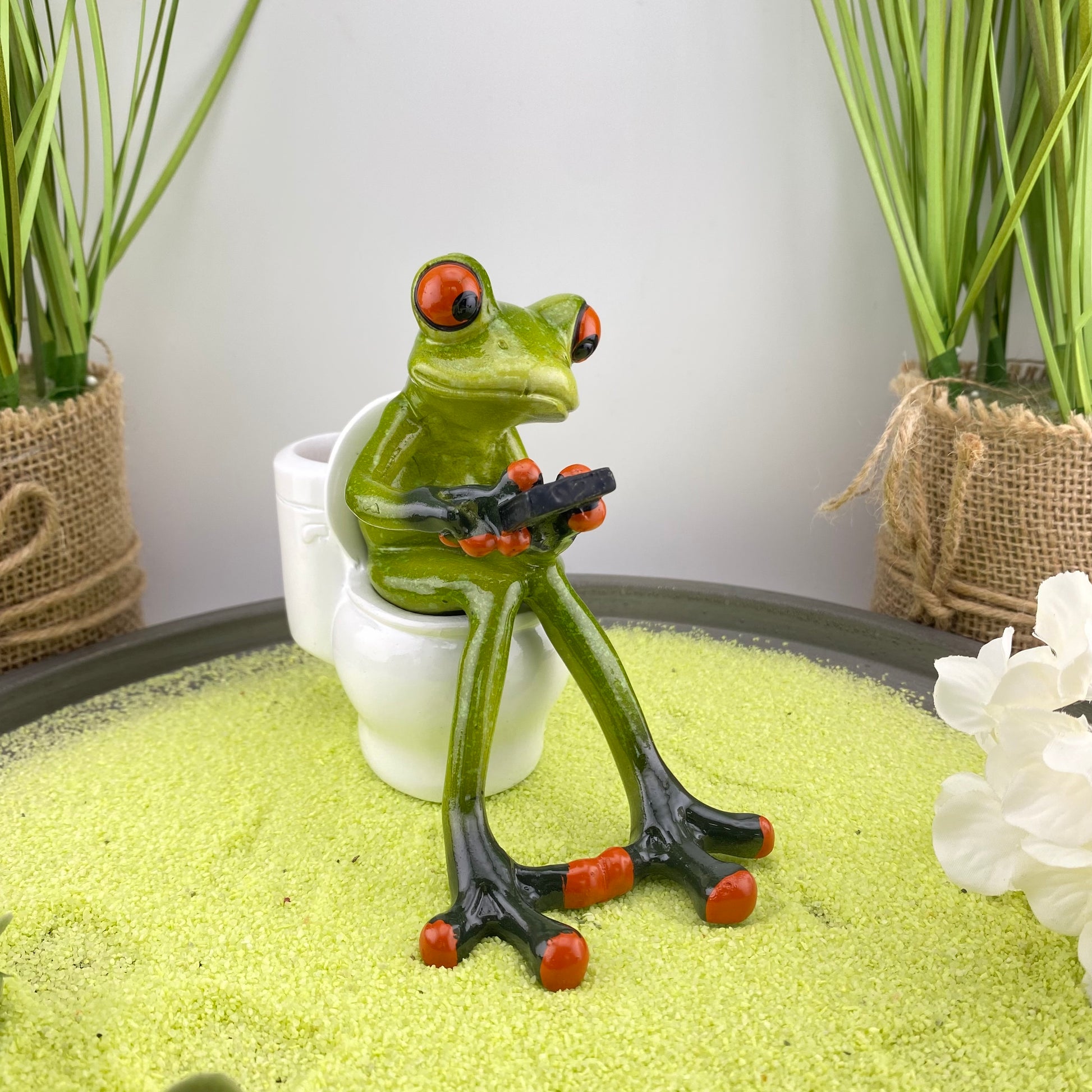 Eine Dekofigur im Ambiente, die einen Frosch auf Toilette abbildet. Der Deko Frosch ist einer ganz alltäglichen Situation mit seinem Smartphone in der Hand dargestellt.