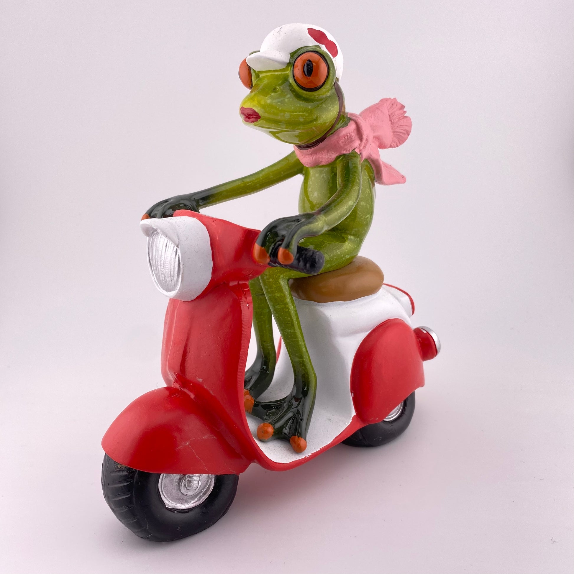Eine Dekofigur vor weißem Hintergrund, die einen weiblichen Frosch auf einem rotem Roller abbildet.