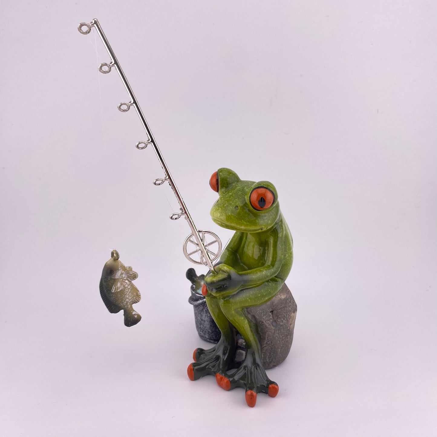 Eine Dekofigur vor weißem Hintergrund, die einen Frosch Mann beim Angeln abbildet. Der Deko Frosch sitzt gemütlich und schaut seinen geangeleten Fisch am Haken seiner Angel an.