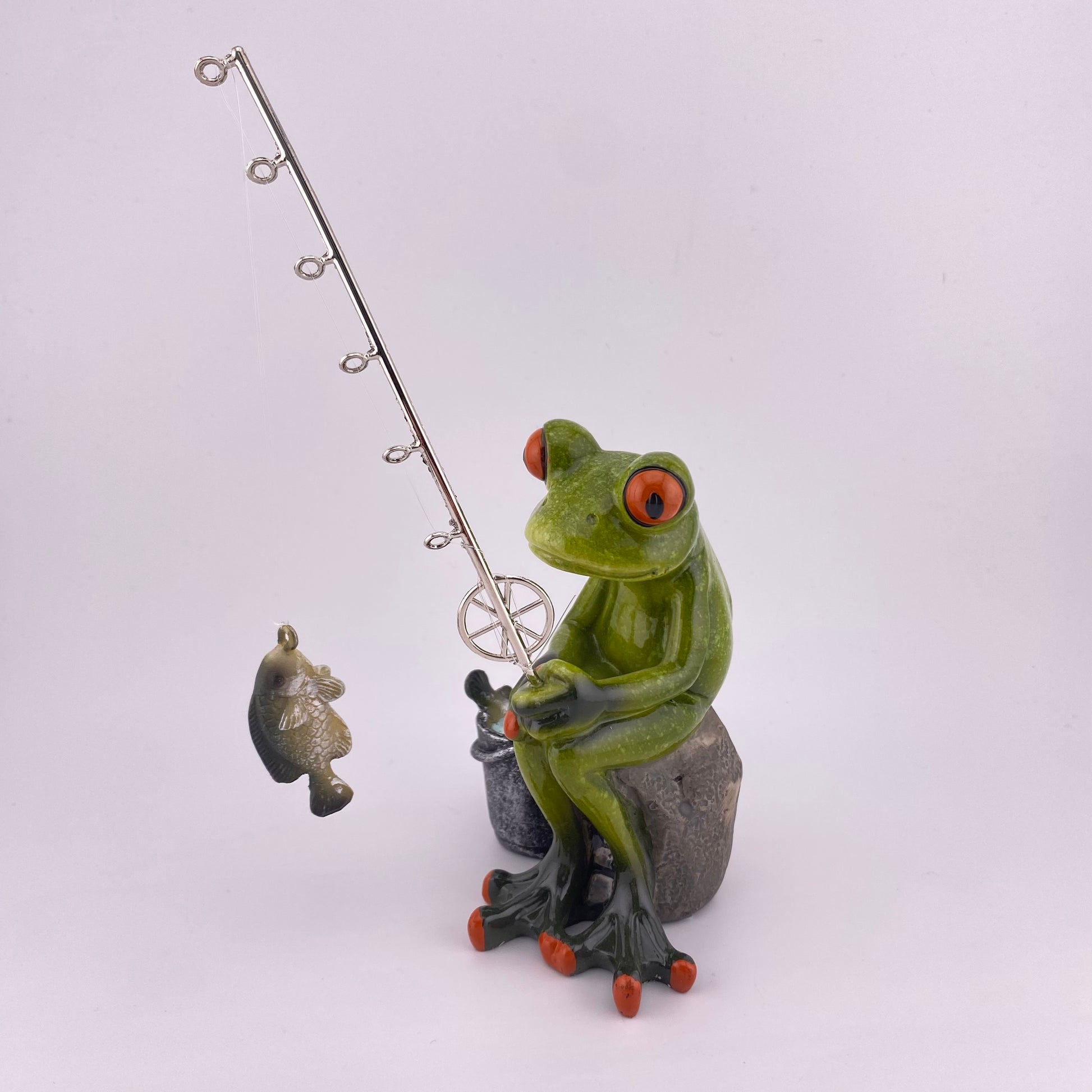 Eine Dekofigur vor weißem Hintergrund, die einen Frosch Mann beim Angeln abbildet. Der Deko Frosch sitzt gemütlich und schaut seinen geangeleten Fisch am Haken seiner Angel an.