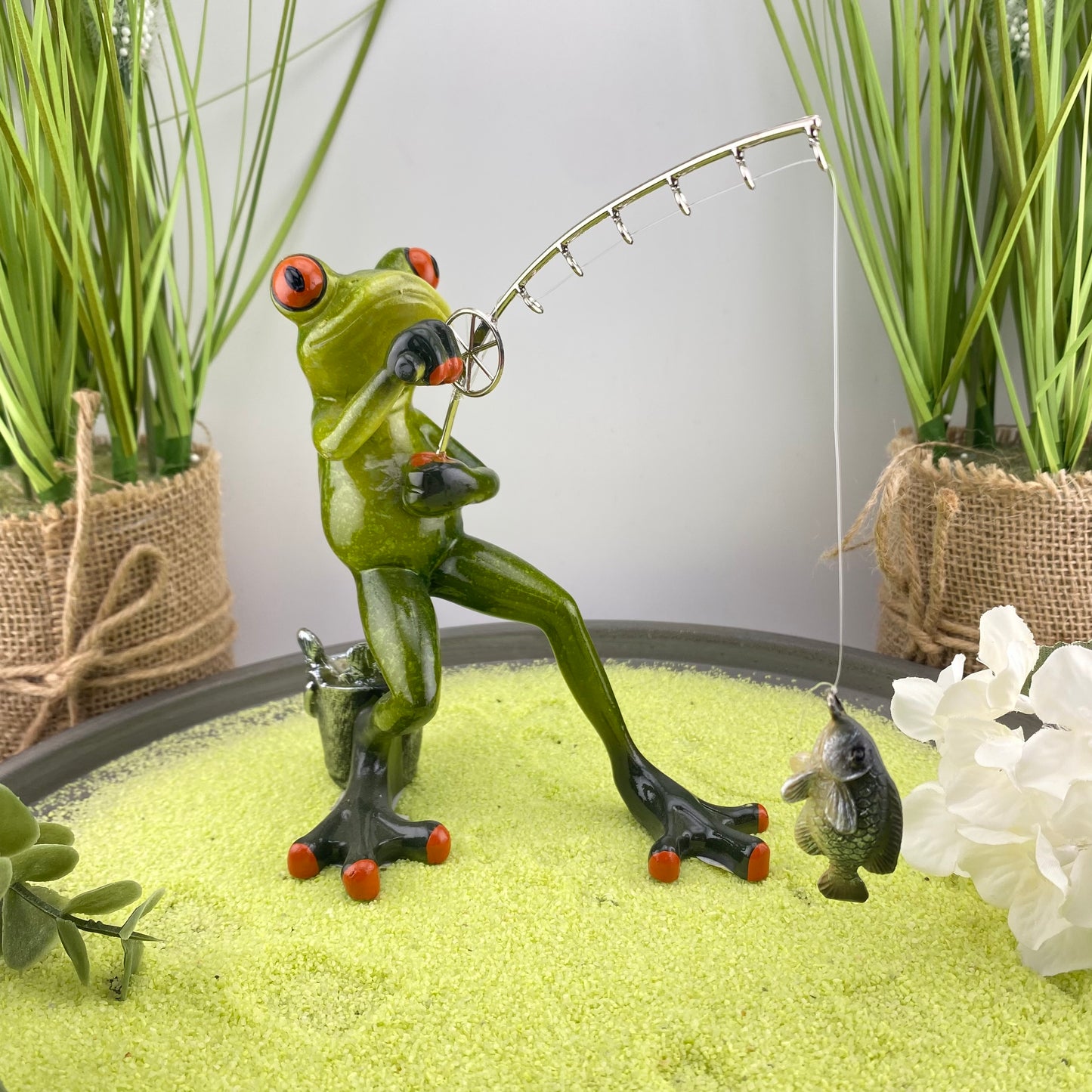 Eine Dekofigur im Ambiente, die einen Frosch Mann beim Angeln abbildet. Der Deko Frosch steht und angelt mit all seiner Kraft einen Fisch, der am Haken seiner Angel hängt. 